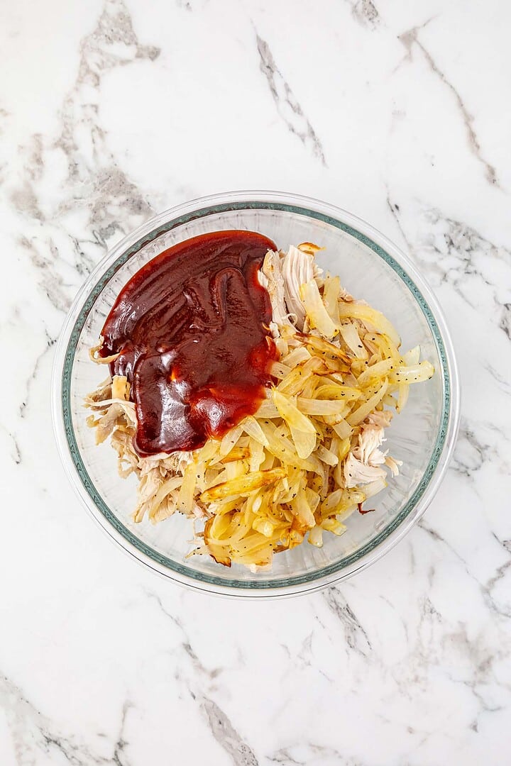 A glass bowl with shredded chicken, cooked onions, and barbecue sauce on a marble countertop—perfect for making Easy Chicken Sliders.