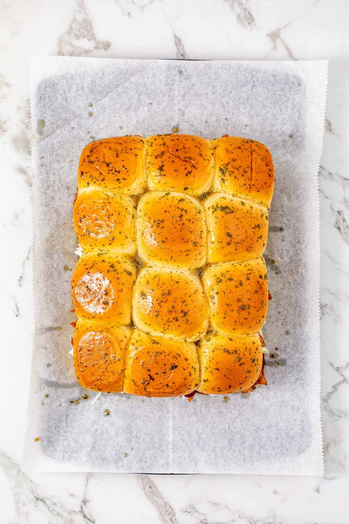 Twelve golden-brown Easy Chicken Sliders topped with herbs rest on parchment paper, arranged neatly on a marble surface.