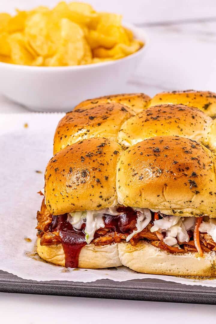 BBQ Chicken Sliders with coleslaw are served on a baking tray, with a bowl of potato chips in the background.