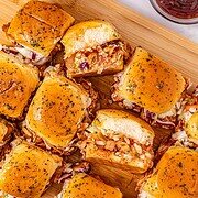 Easy BBQ Chicken Sliders with tangy coleslaw are served on a wooden board, accompanied by crispy chips and a bowl of barbecue sauce.