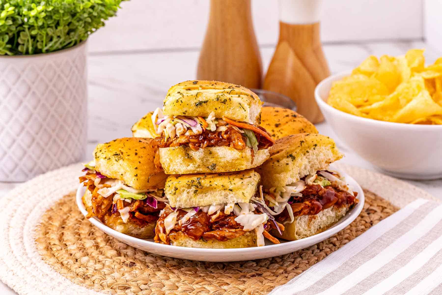 A plate of BBQ chicken sliders piled with coleslaw sits beside a bowl of potato chips on a woven placemat, making for an easy chicken sliders meal.