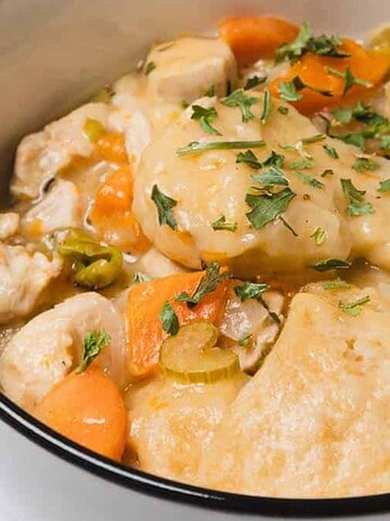 A bowl of chicken and dumplings with carrots, celery, and herbs, with a spoon inside.