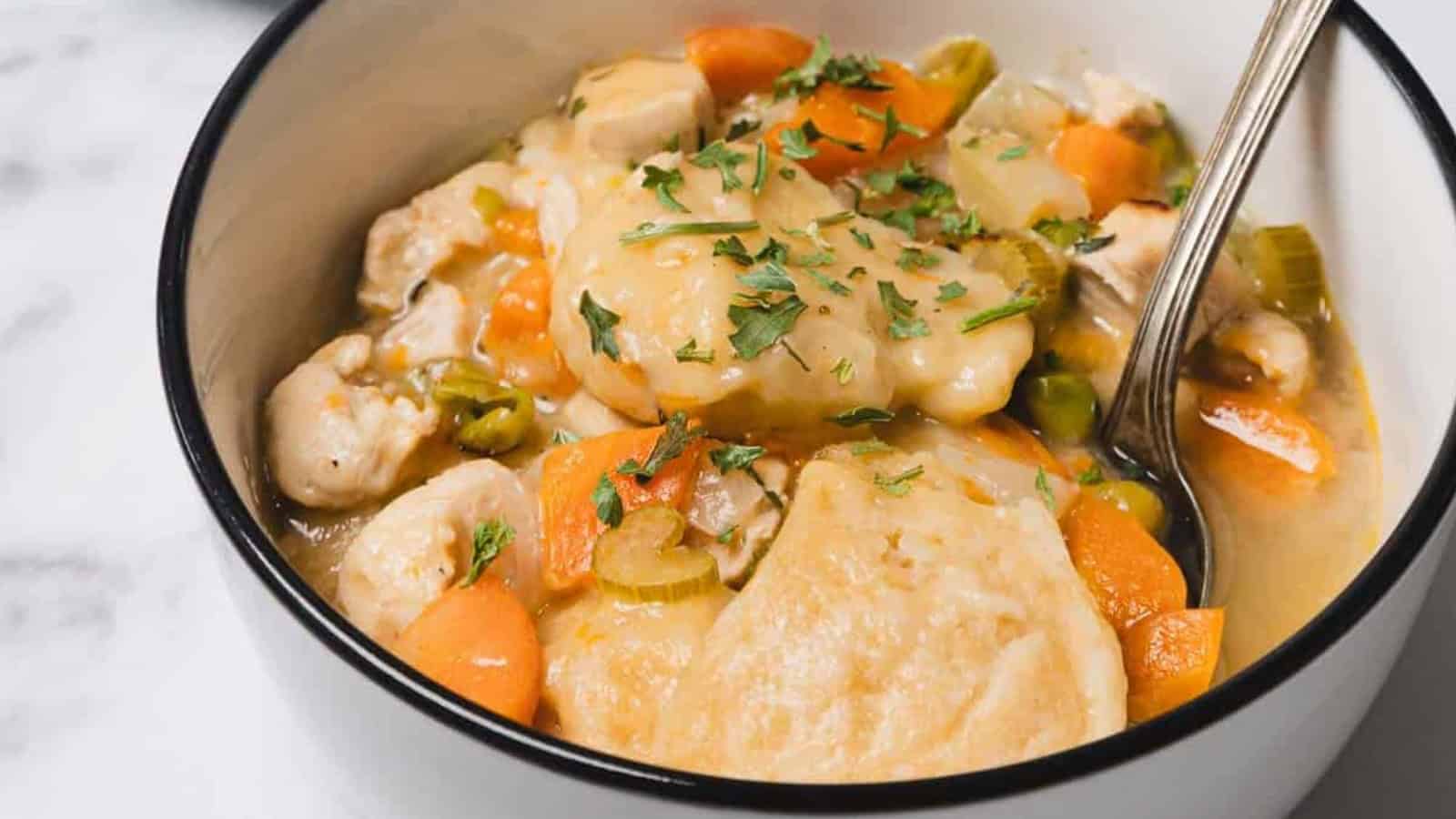 A bowl of chicken and dumplings soup with carrots, celery, and herbs, with a spoon inside.