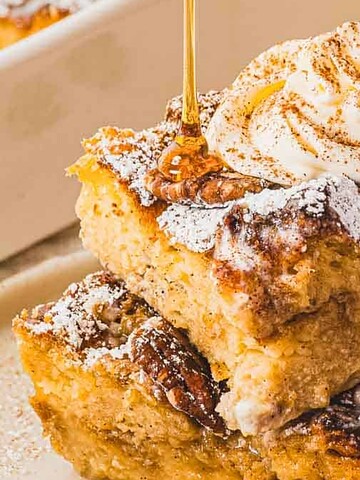 Two slices of bread pudding stacked, topped with whipped cream, cinnamon, and powdered sugar on a plate.