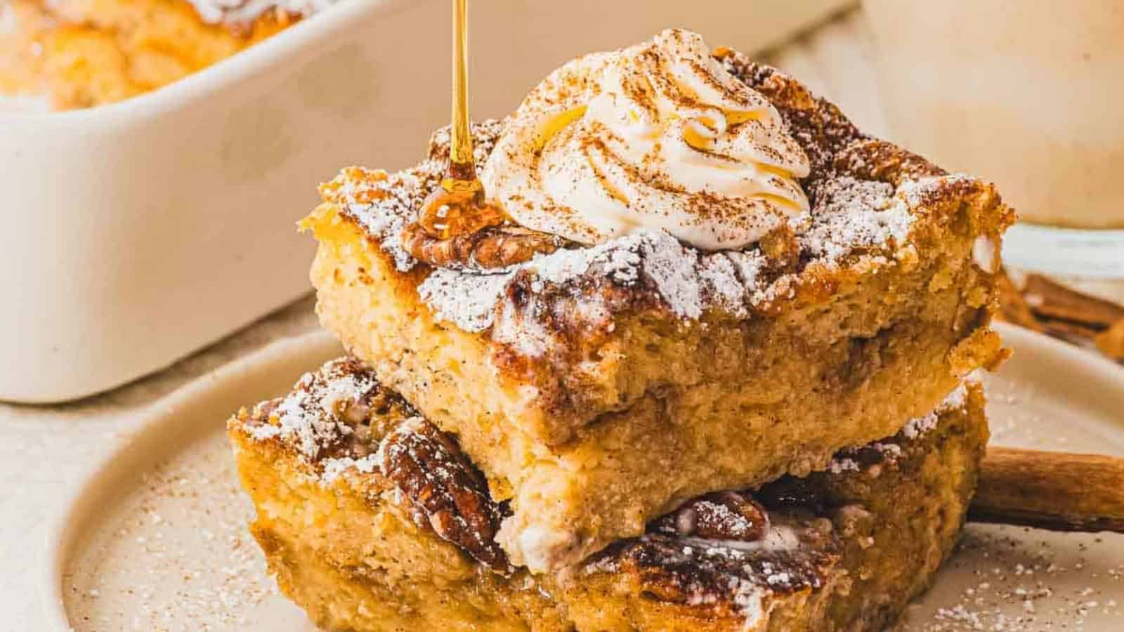 Two slices of bread pudding stacked, topped with whipped cream, cinnamon, and powdered sugar on a plate.