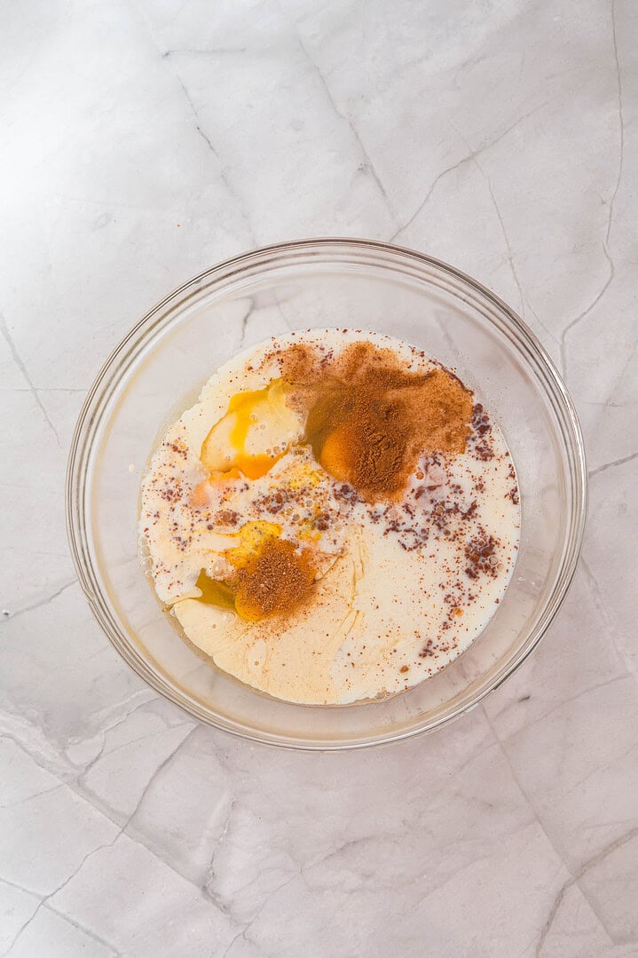 A glass bowl with eggs, milk, and spices on a marble countertop, ready to be mixed for a delicious Eggnog French Toast casserole.