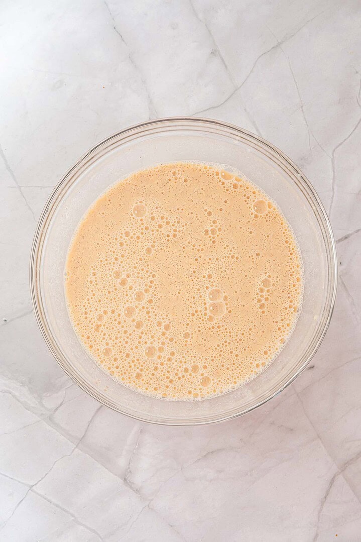 A glass bowl filled with a frothy, light orange liquid on a white marble surface&mdash;perfect for making Eggnog French Toast or your favorite Eggnog Casserole.