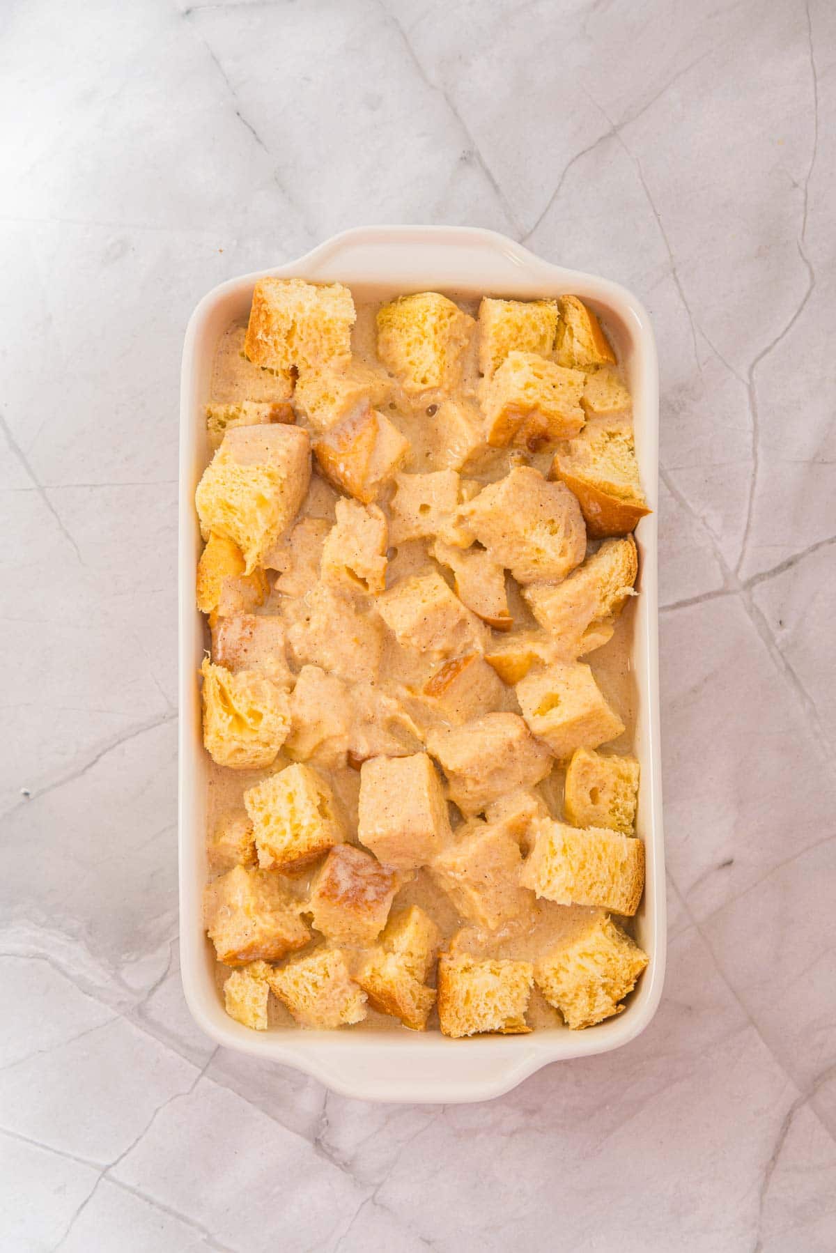 Chunks of bread soaked in a creamy eggnog mixture fill a white baking dish on a marble countertop, creating a delicious French Toast casserole.