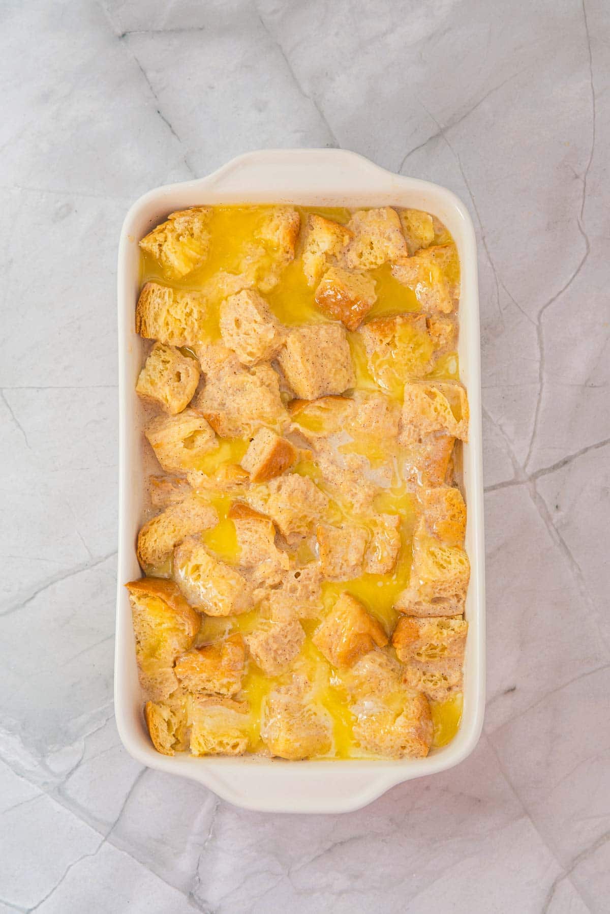 Cubed bread soaked in a rich egg mixture for Eggnog French Toast casserole, ready to bake in a white dish on a marble surface.