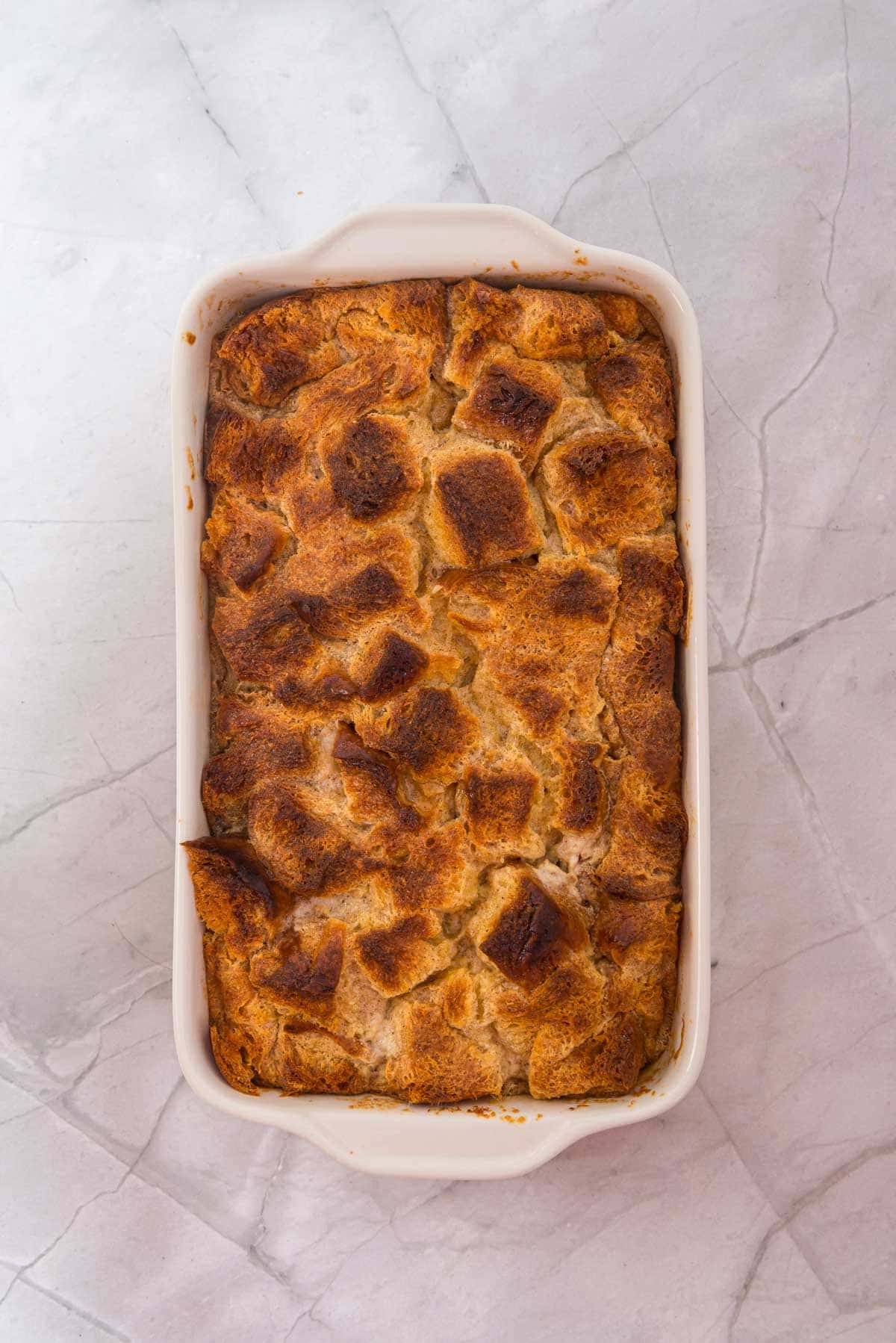 A delicious Eggnog French Toast casserole baked in a rectangular white dish sits on a light marble surface.