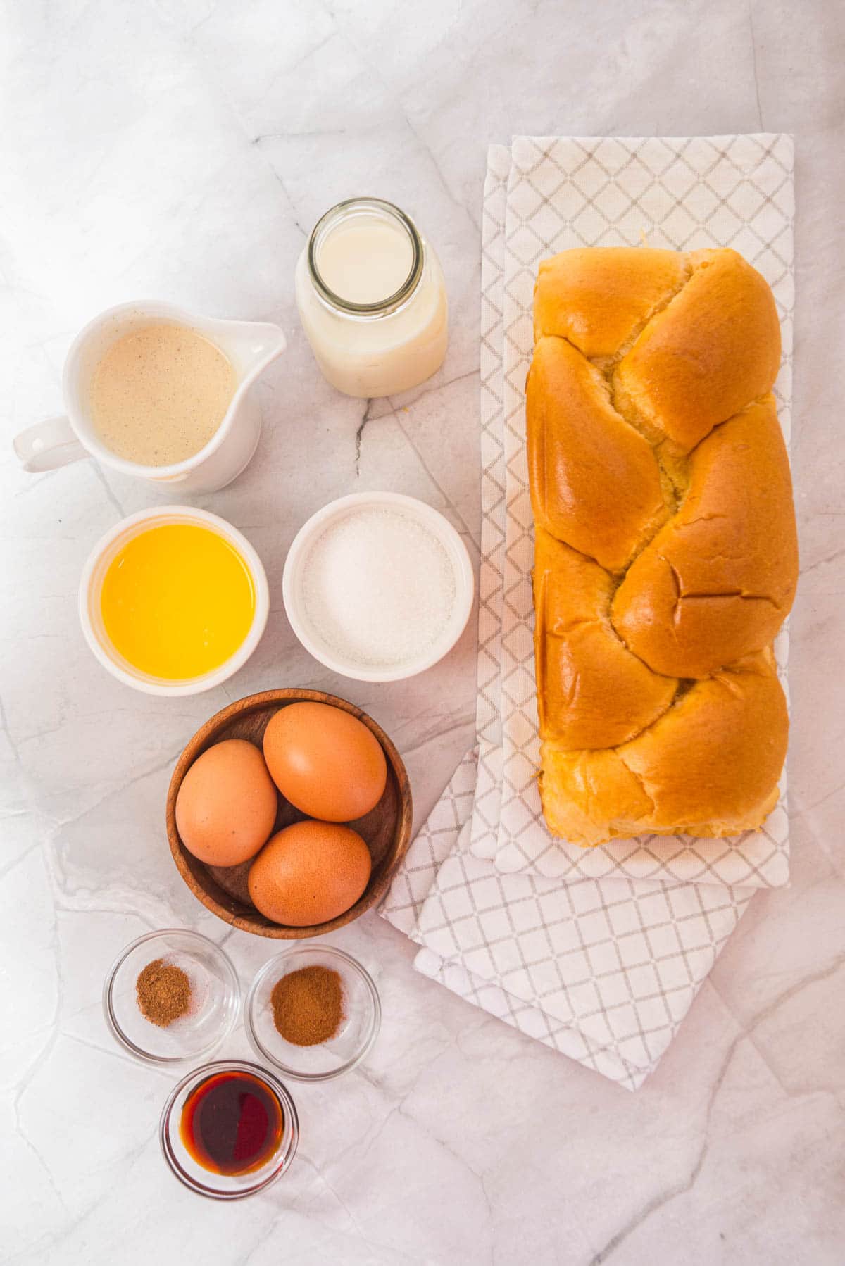 A loaf of bread, eggs, milk, melted butter, sugar, and spices arranged on a marble surface&mdash;perfect ingredients for a festive Eggnog French Toast Casserole.