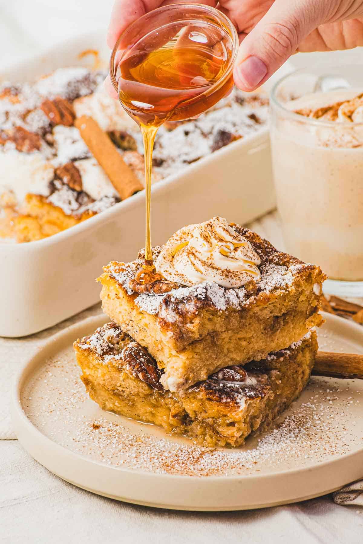 A hand pours syrup over two pieces of Eggnog French Toast topped with whipped cream and powdered sugar.