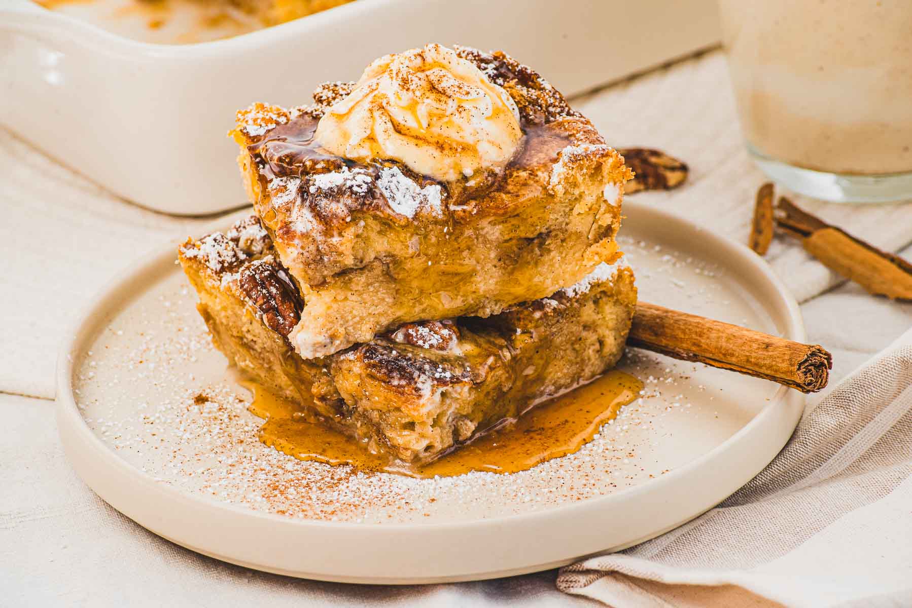 Two pieces of Eggnog French Toast topped with whipped cream, powdered sugar, and syrup on a plate with a cinnamon stick&mdash;just like a festive bread pudding treat.
