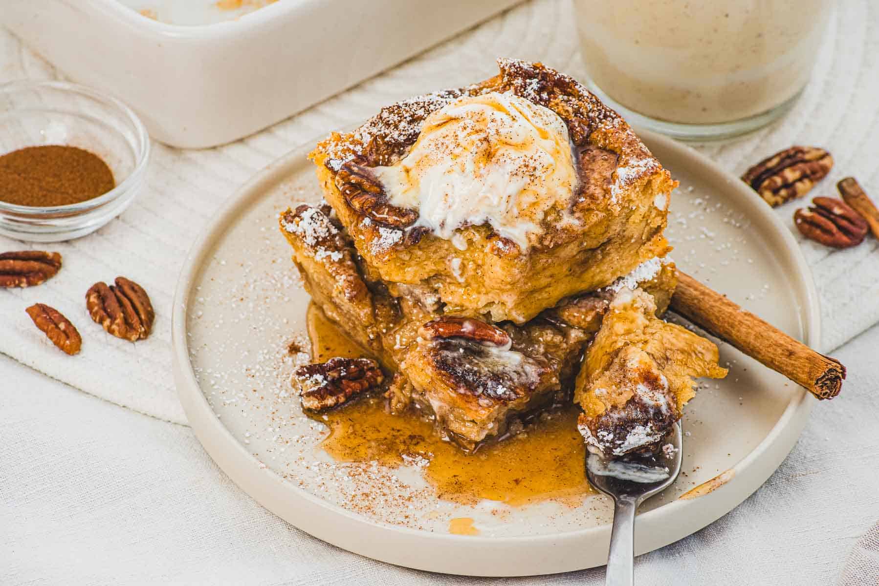 A plate of Eggnog French Toast Casserole topped with ice cream, pecans, powdered sugar, and syrup, garnished with a cinnamon stick.
