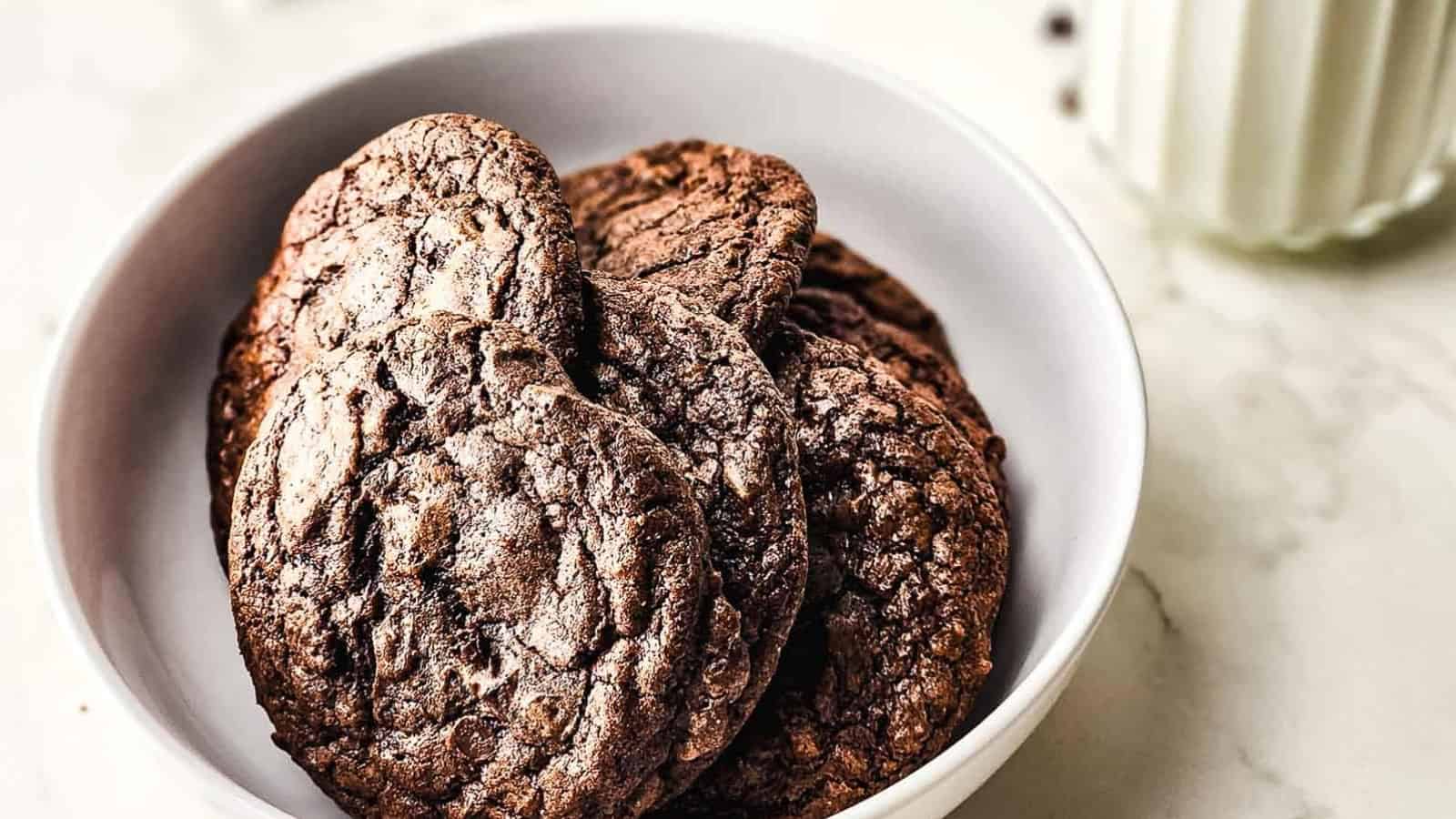 A white bowl filled with several chocolate cookies, with a glass of milk in the background.