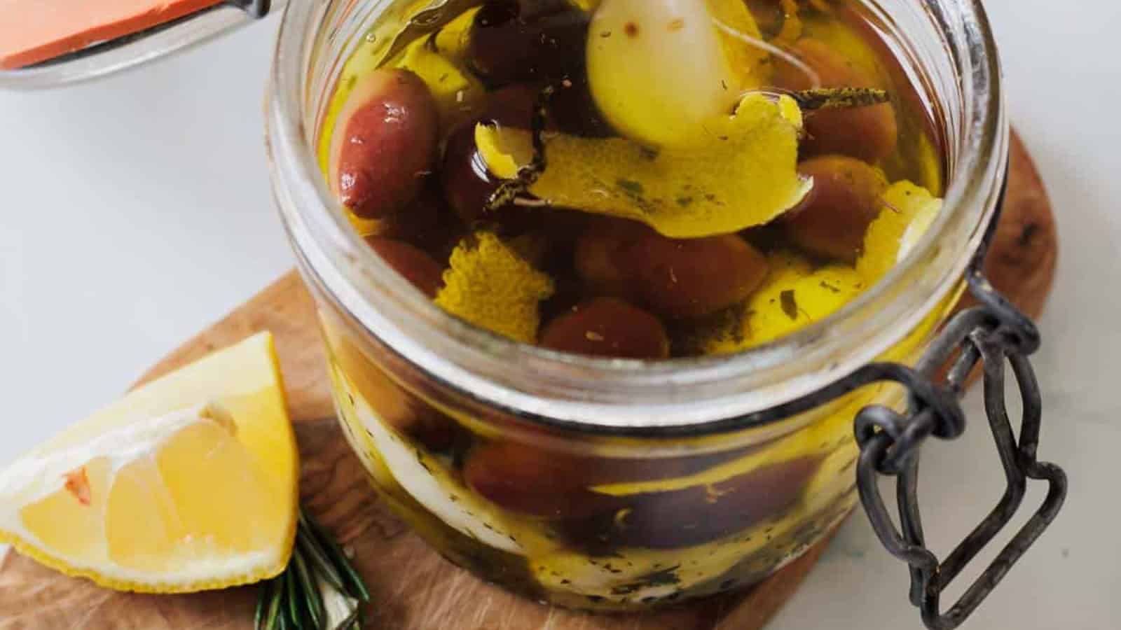 A jar of marinated olives with herbs and lemon peel, placed on a wooden board next to lemon wedges.