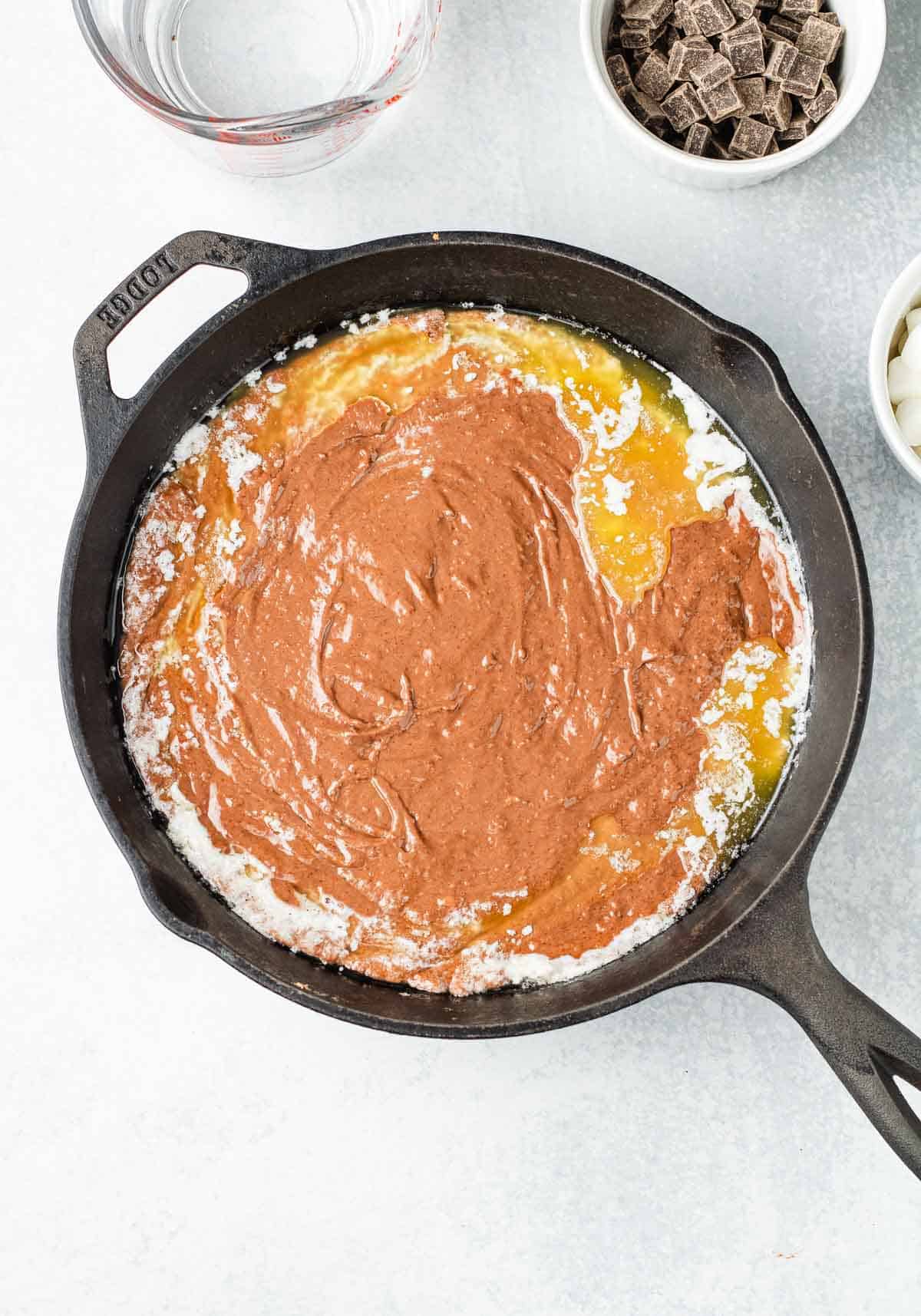 A cast iron skillet with brownie batter swirled into melted butter and sugar, surrounded by baking ingredients&mdash;a tempting setup for a decadent hot chocolate cobbler recipe.