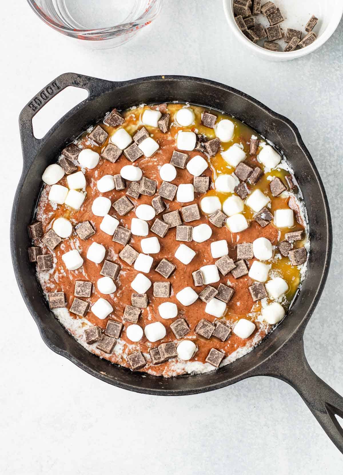 A cast iron skillet filled with chocolate chunks and mini marshmallows on a batter mixture, perfect for creating a decadent Hot Chocolate Cobbler.