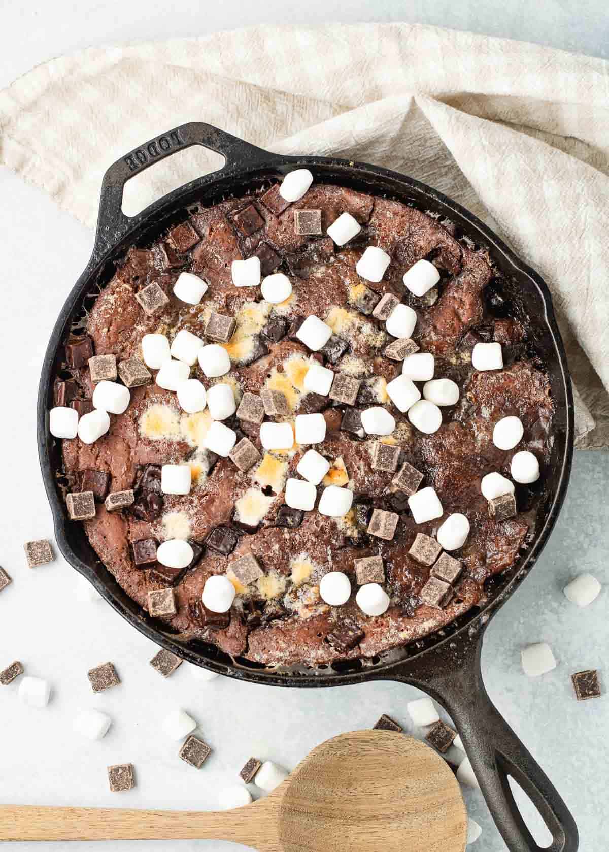 A hot chocolate dessert skillet brownie topped with marshmallows and chocolate chunks, with a wooden spoon and napkin nearby.