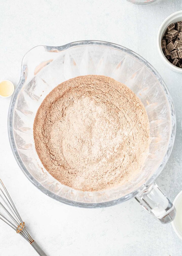 Large glass bowl with cocoa powder and flour mixture on a white surface, surrounded by baking tools&mdash;perfect for starting your Hot Chocolate Cobbler or any cozy Hot Chocolate Dessert.