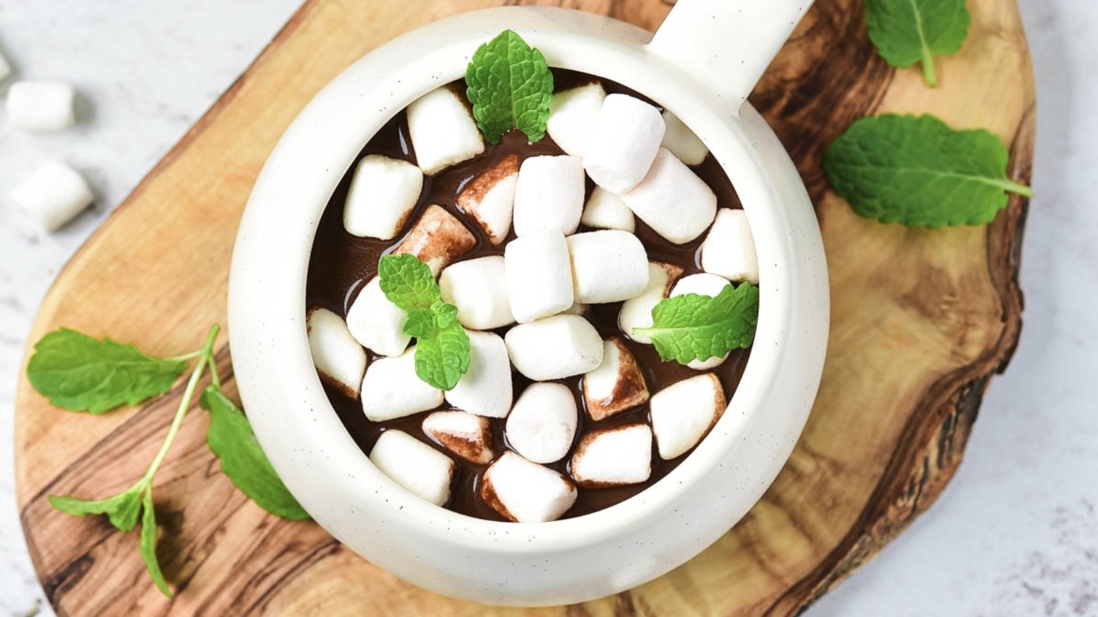 A mug of hot chocolate topped with mini marshmallows and mint leaves on a wooden board.