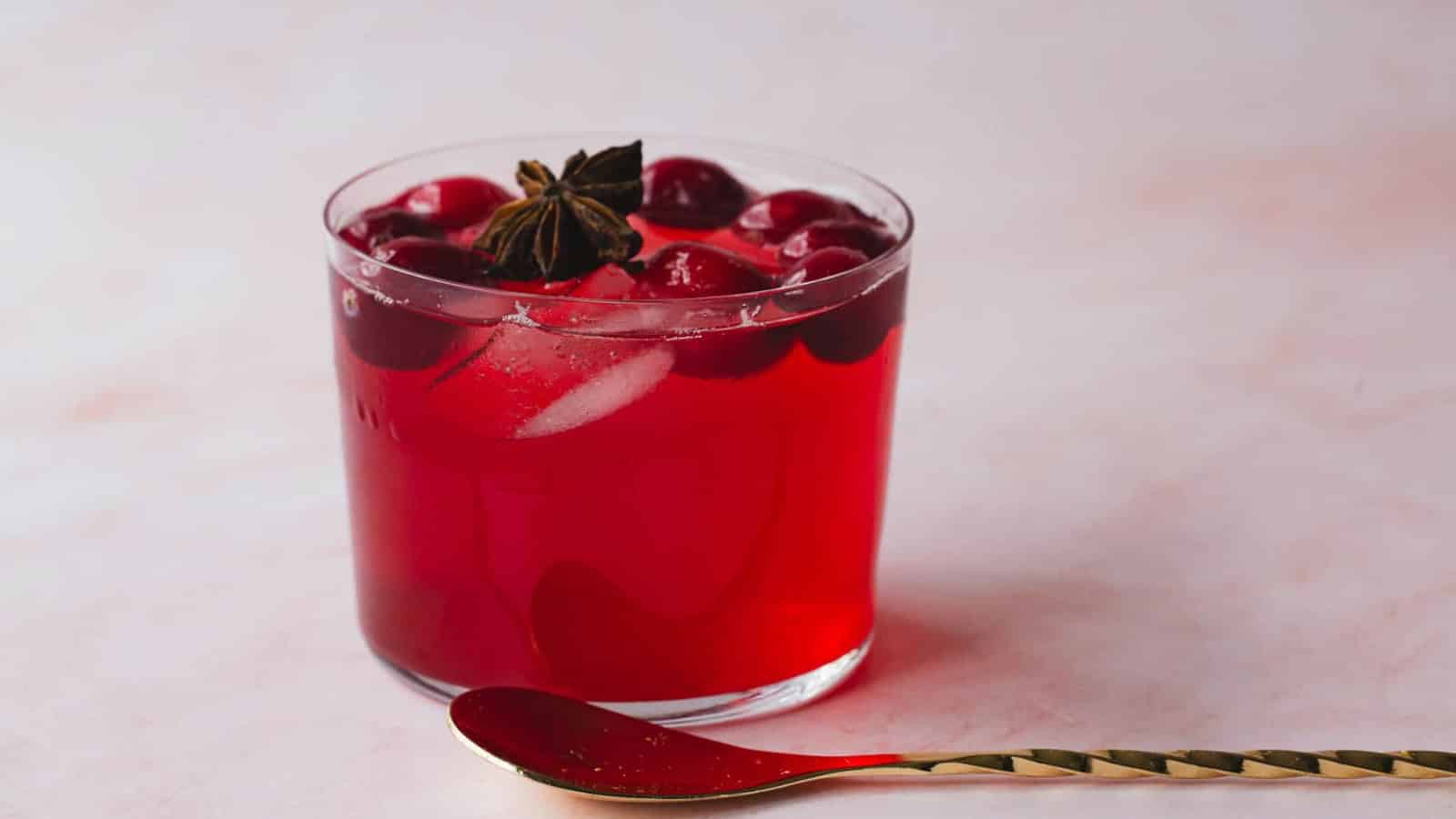 A glass of red iced drink with cherries and star anise, next to a gold spoon on a light surface.