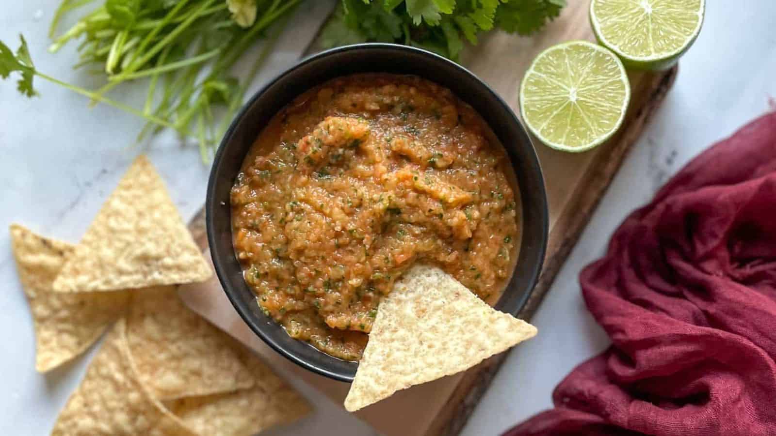 A bowl of pineapple habanero salsa surrounded by cilantro, limes, and a red linen sit on a wooden cutting board.