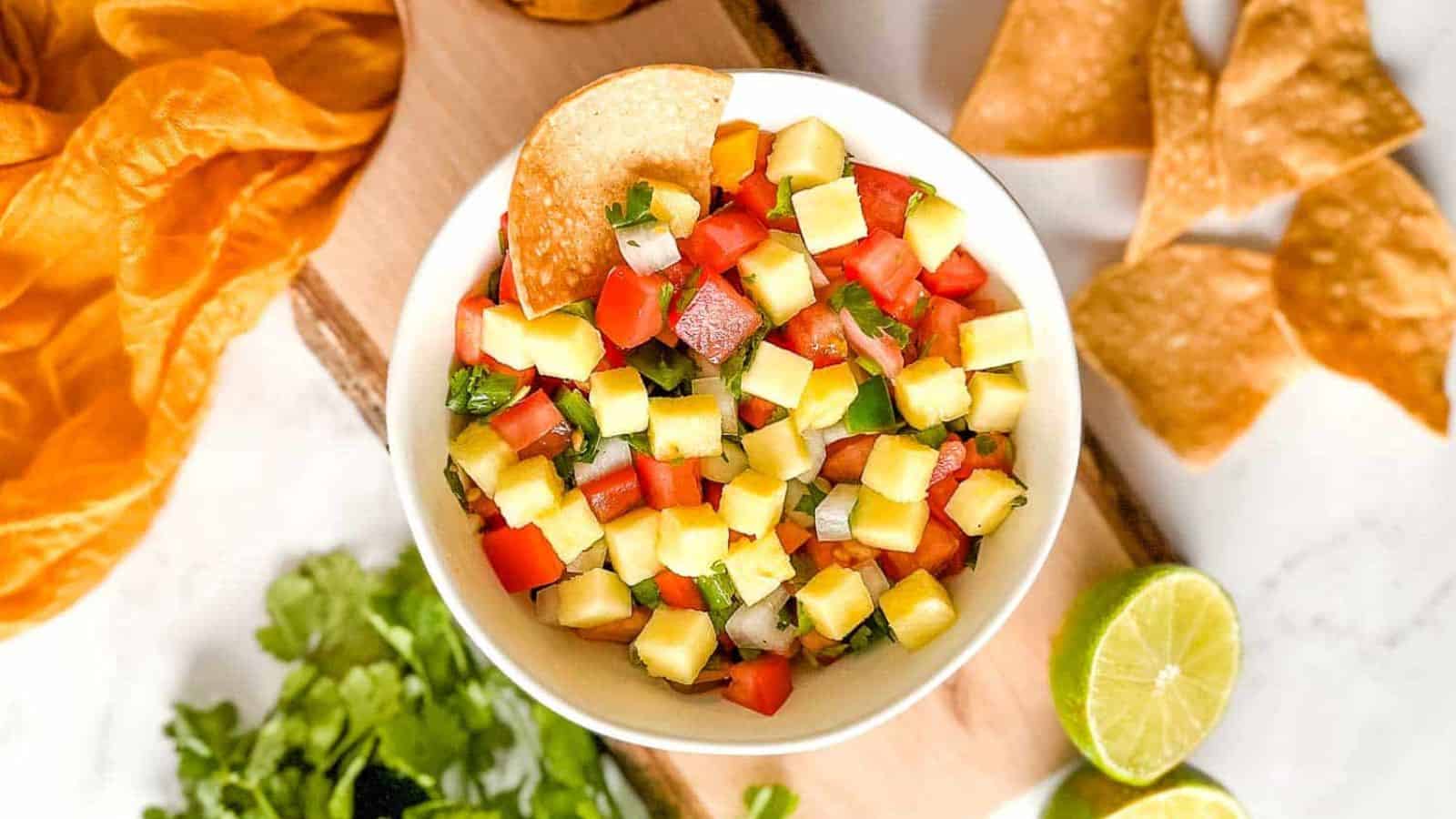 Pineapple pico de gallo in a black bowl surrounded by limes and a yellow linen.