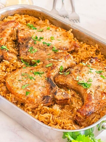 Four cooked pork chops served over seasoned rice in a large silver baking dish, garnished with fresh herbs.