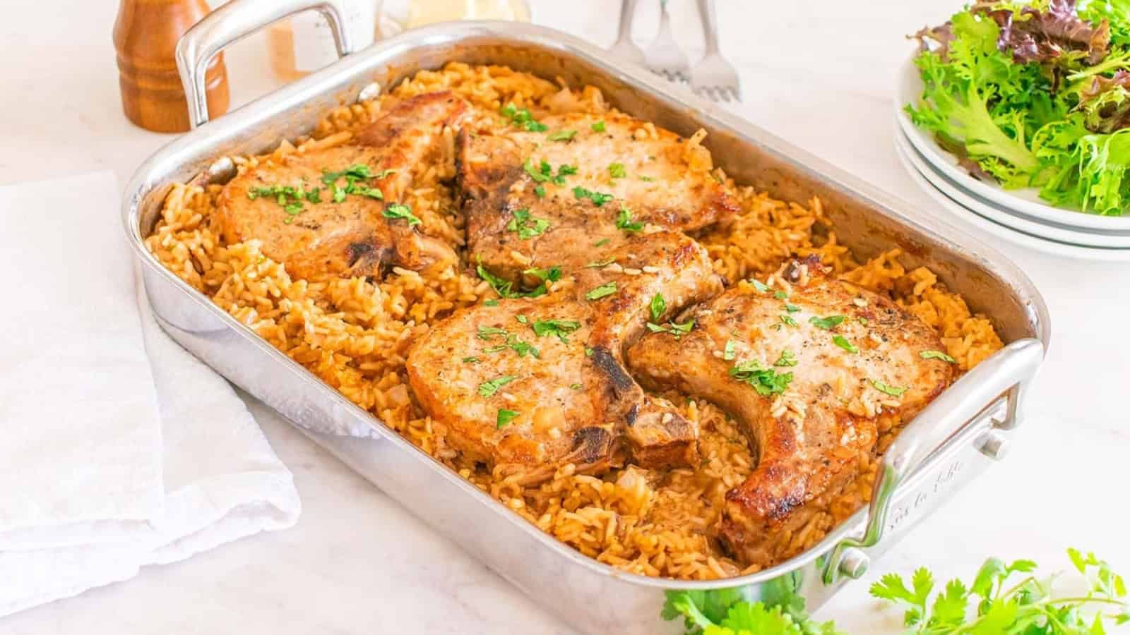 A baking dish with four cooked pork chops on top of seasoned rice, garnished with chopped herbs. Plates and a salad are in the background.
