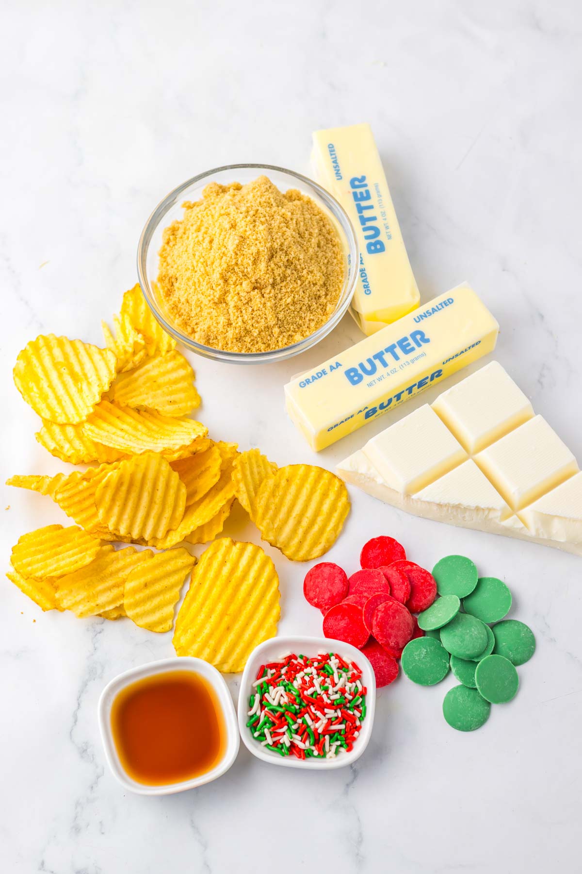 Potato Chip Christmas Crack features potato chips, graham crumbs, butter, white chocolate, candy melts, sprinkles, and vanilla&mdash;all spread out on a white surface for a festive treat.