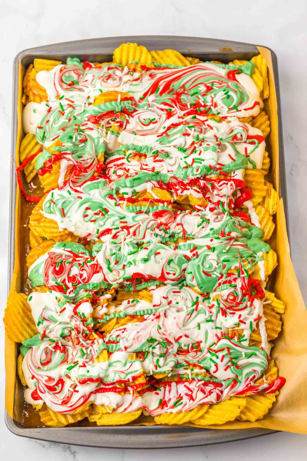 Wavy potato chips topped with red, green, and white swirled frosting and sprinkles create a festive Potato Chip Christmas Crack on a baking tray.
