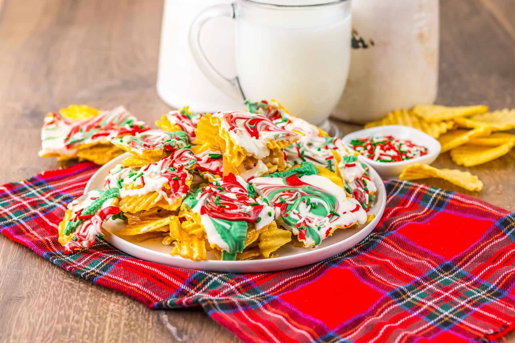 A plate of wavy potato chips drizzled in festive white, red, and green&mdash;like a Christmas Crack recipe&mdash;sits on a plaid cloth next to a glass of milk.