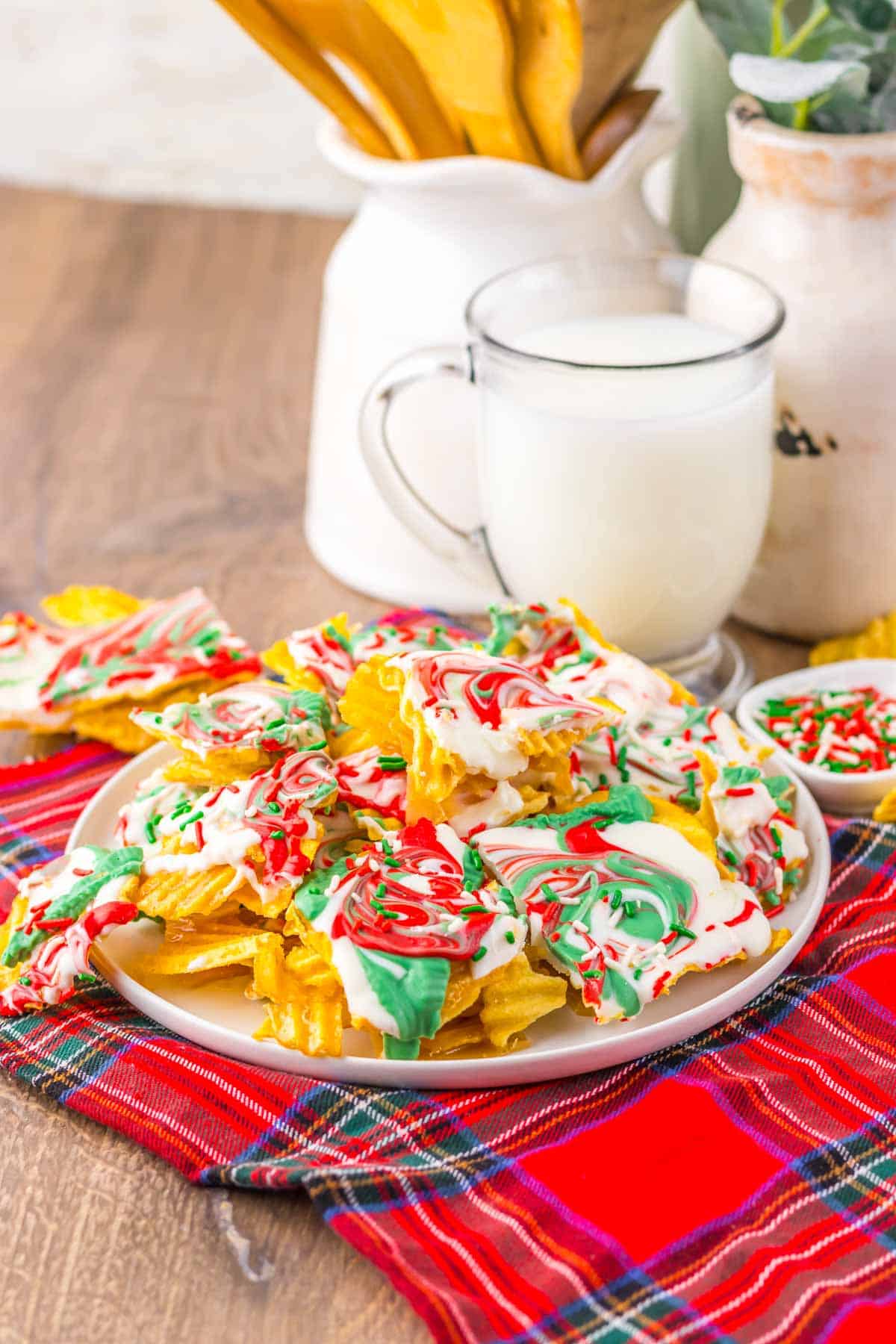 Plate of potato chips drizzled with red, white, and green chocolate&mdash;like a festive Christmas Crack recipe&mdash;sits next to a glass of milk on a plaid cloth.