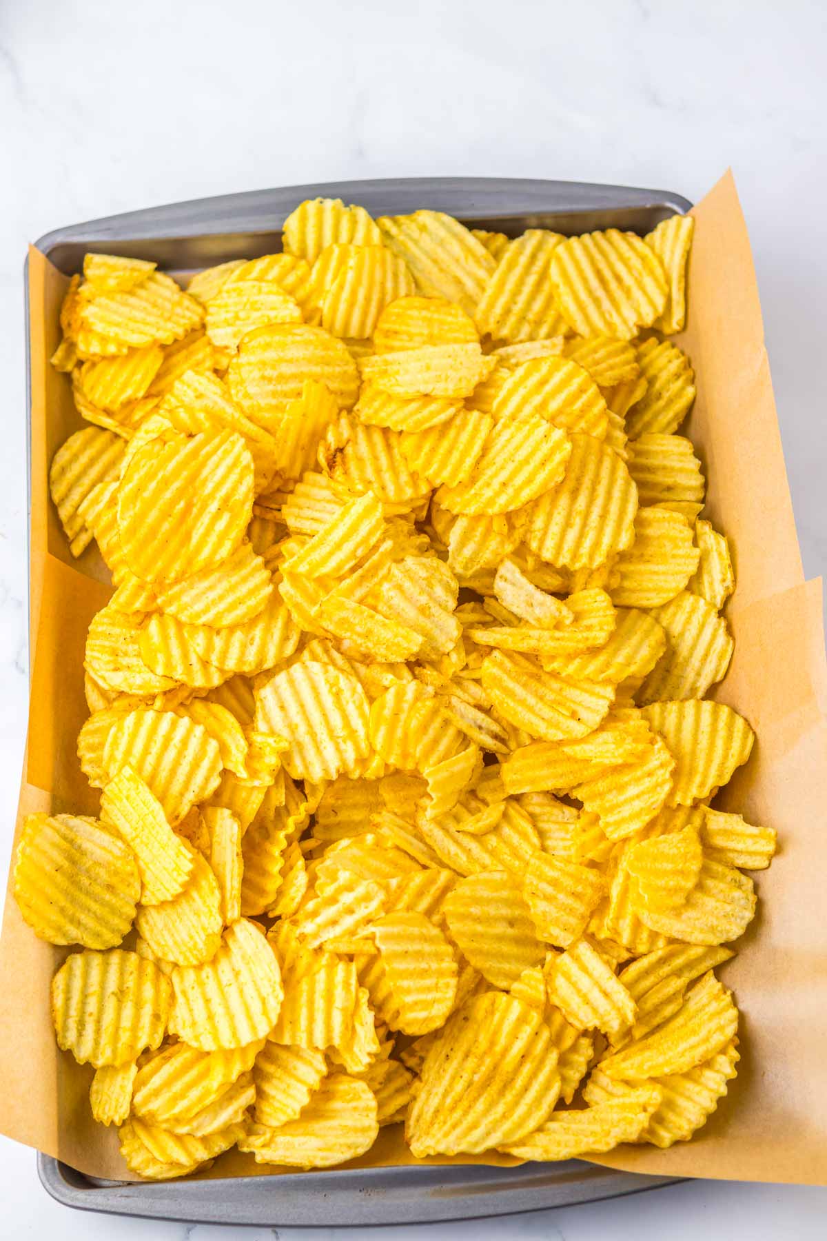 A baking tray lined with parchment paper, filled with wavy, ridged potato chips&mdash;perfect for trying a new potato chip recipe or adding a twist to your Christmas Crack treats.