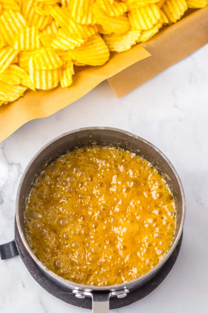 Wavy potato chips next to a pot of bubbling caramel sauce on a marble surface inspire the perfect Potato Chip Christmas Crack recipe.