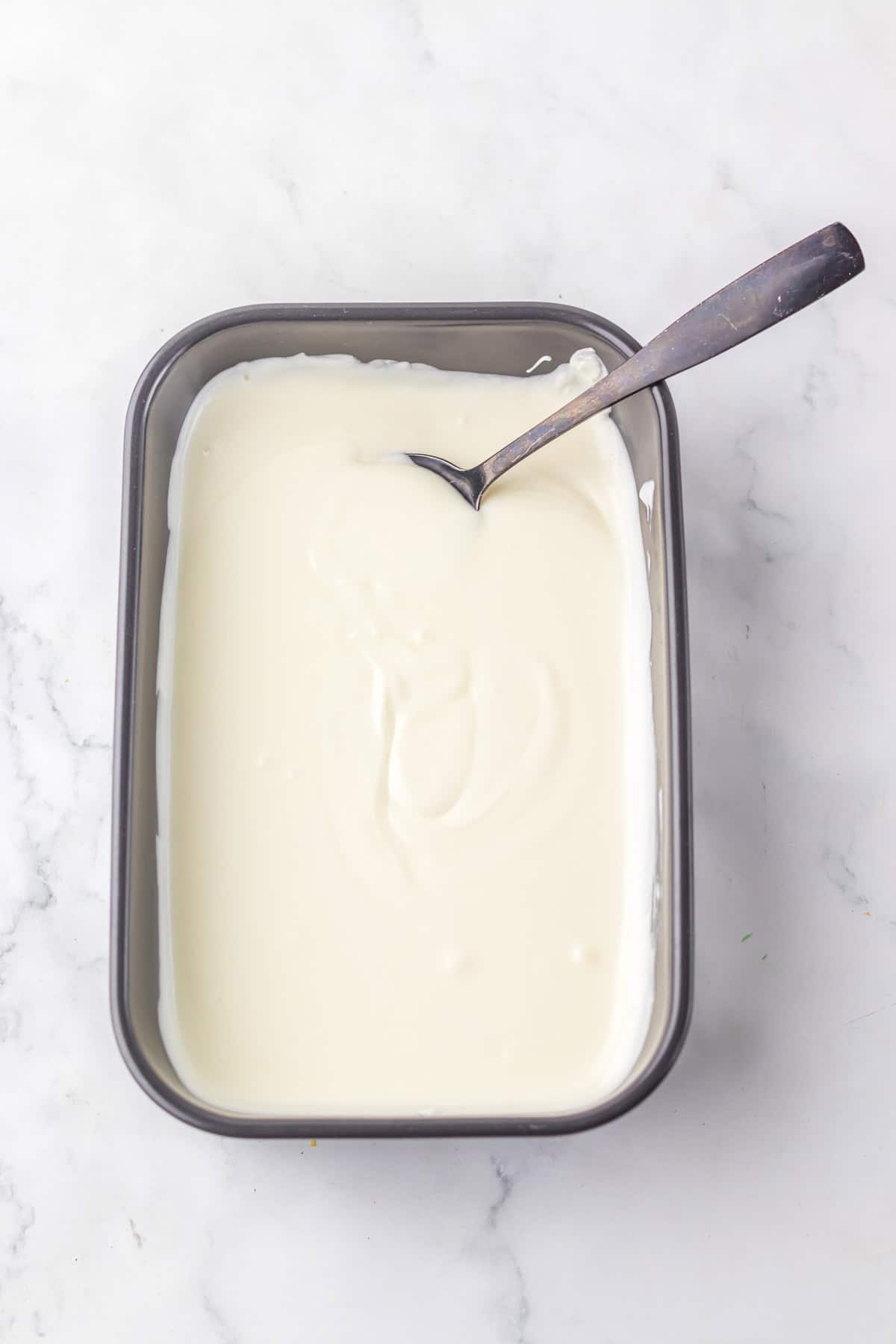 A rectangular pan filled with white yogurt and a spoon resting inside on a marble surface, reminiscent of the layered look of Potato Chip Christmas Crack.