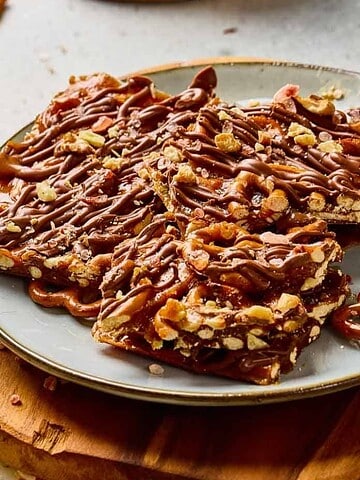 A plate of chocolate-covered pretzel bark sits on a wooden board, surrounded by scattered pretzels.