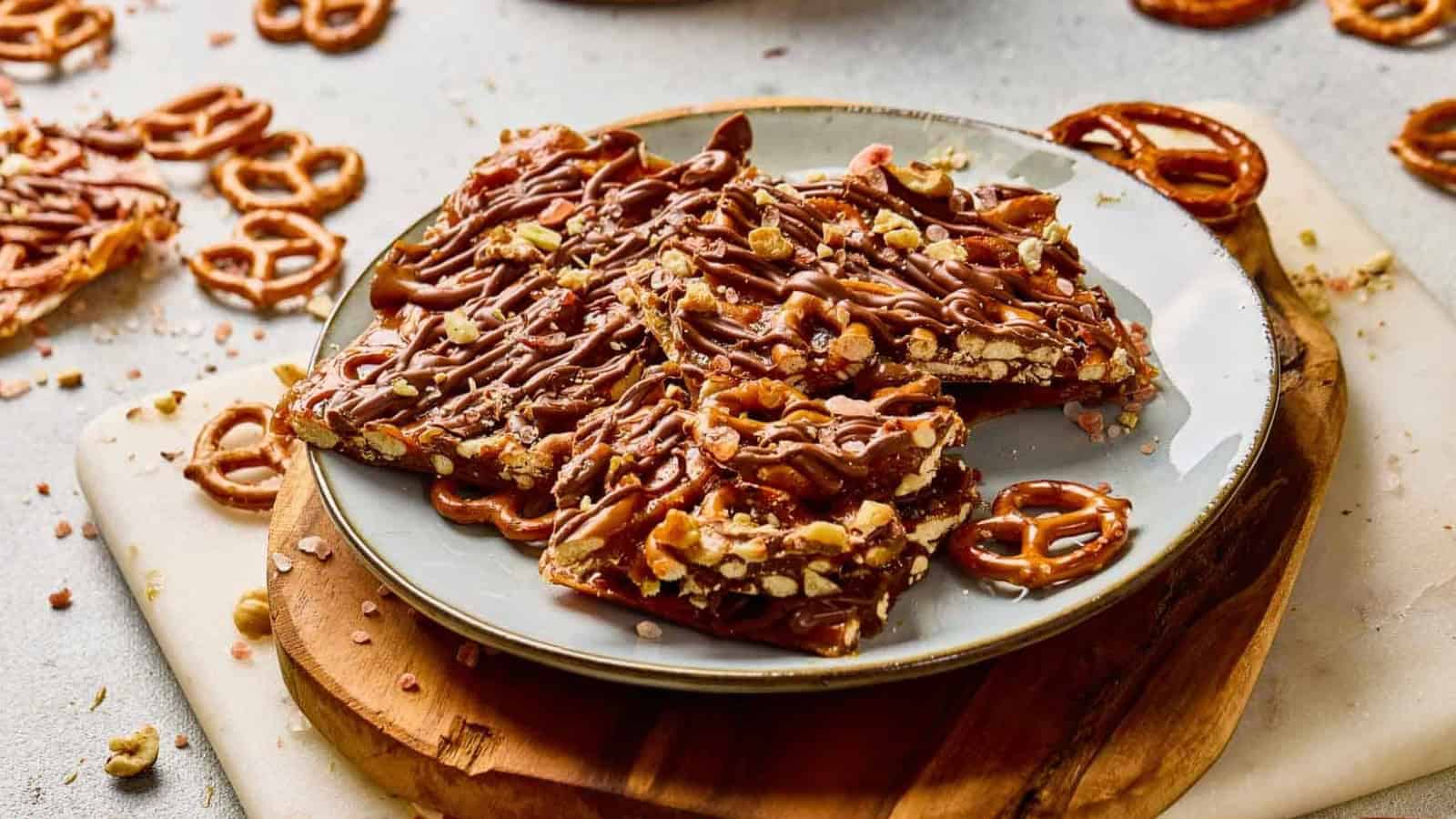 Chocolate-drizzled pretzel toffee pieces on a gray plate, topped with chopped pretzels and nuts.