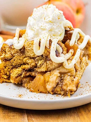 A slice of apple pie with whipped cream and icing is served on a white plate with apples in the background.