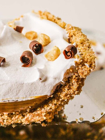 A slice of creamy pie with whipped topping, chocolate curls, and chopped nuts on a crumbly crust.