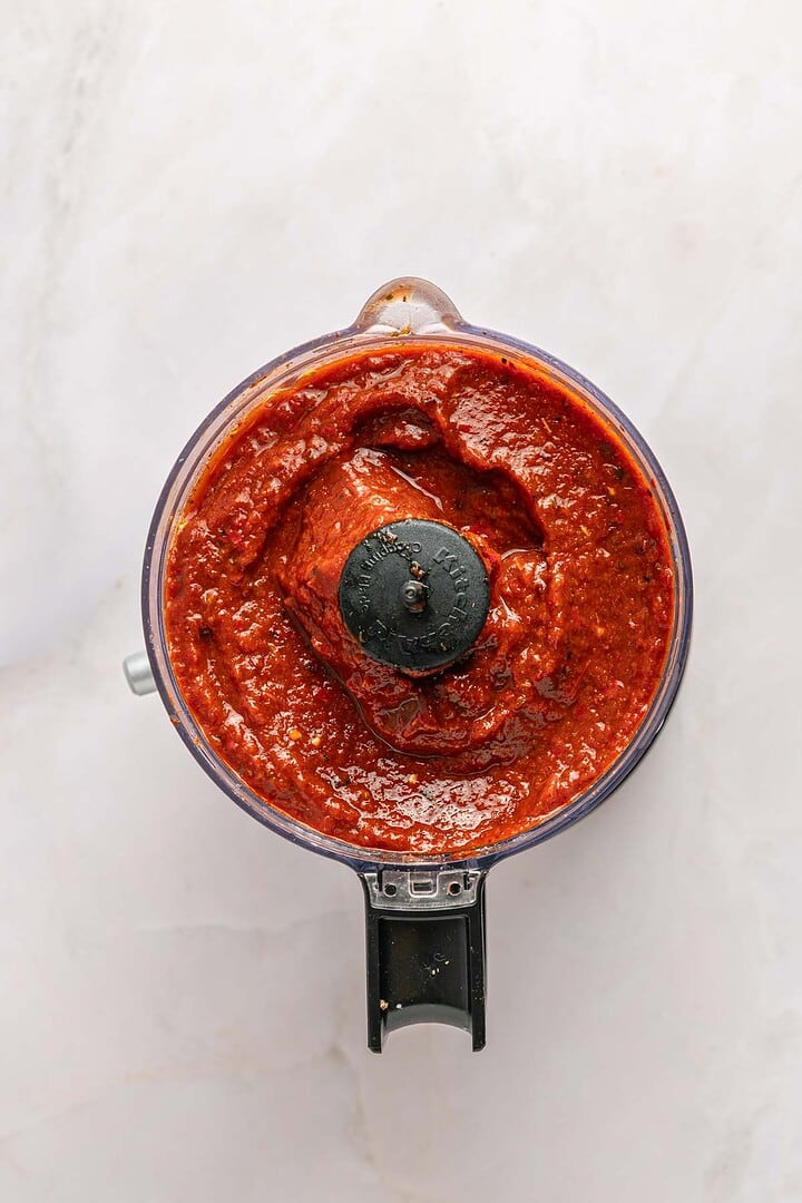 Overhead view of a food processor filled with thick, red Birria sauce on a light countertop&mdash;perfect for making tacos or slow cooker recipes.