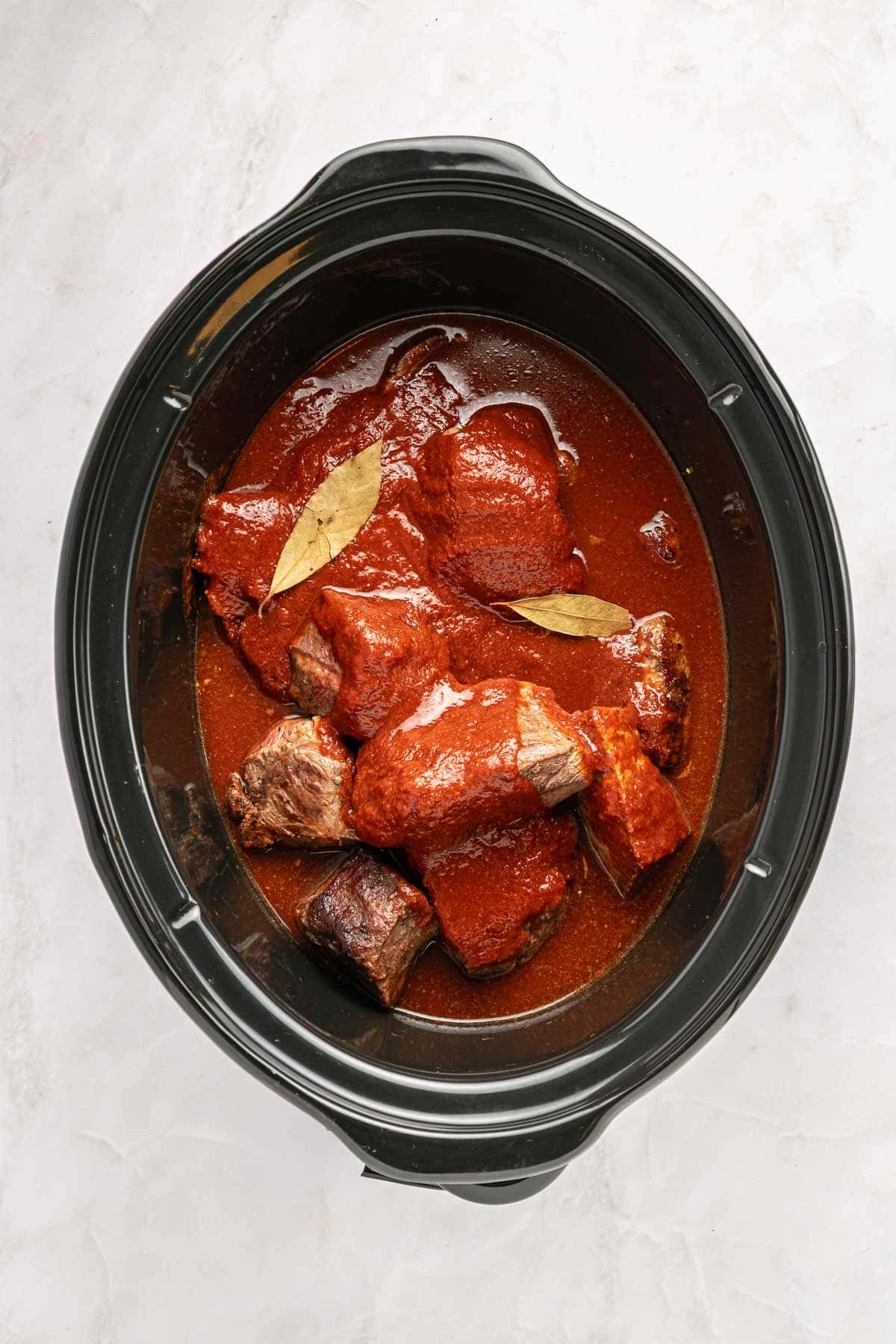 Chunks of beef in red sauce with bay leaves for Slow Cooker Birria simmer in a black slow cooker on a white surface.