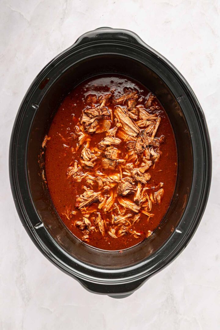 Shredded beef birria in a rich red sauce simmers inside a black slow cooker, perfect for making flavorful tacos on a light marble surface.