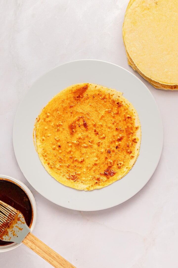 Corn tortilla on a white plate with red sauce spread, perfect for making Slow Cooker Birria Tacos; sauce and brush rest in a bowl beside it.