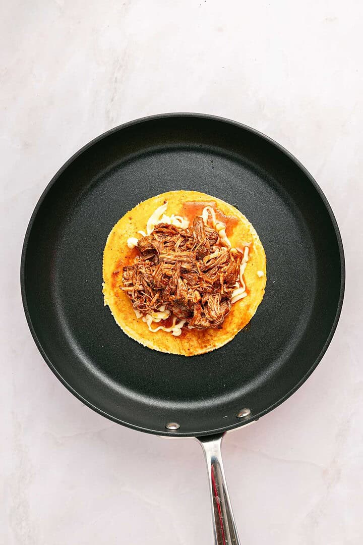 A corn tortilla with shredded beef and rich sauce sits in a black skillet on a light surface, showcasing classic Birria Tacos.