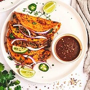 Three slow cooker birria tacos with sliced onions, jalapeños, lime wedges, and a bowl of dipping sauce on a white plate.