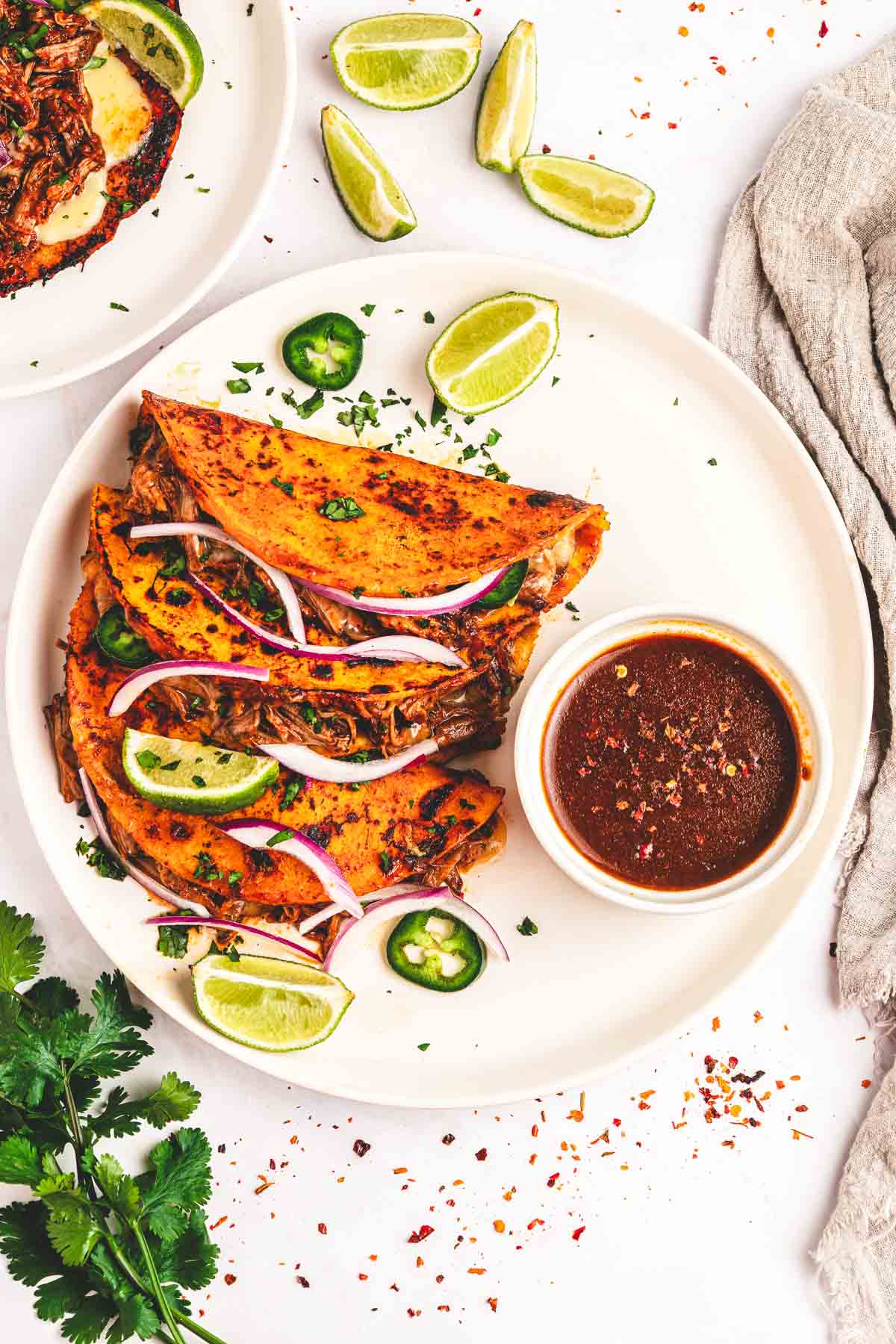 Three birria tacos with shredded meat, red onions, jalape&ntilde;os, lime wedges, and a bowl of dipping sauce&mdash;classic tacos made even better with rich slow cooker birria flavor.