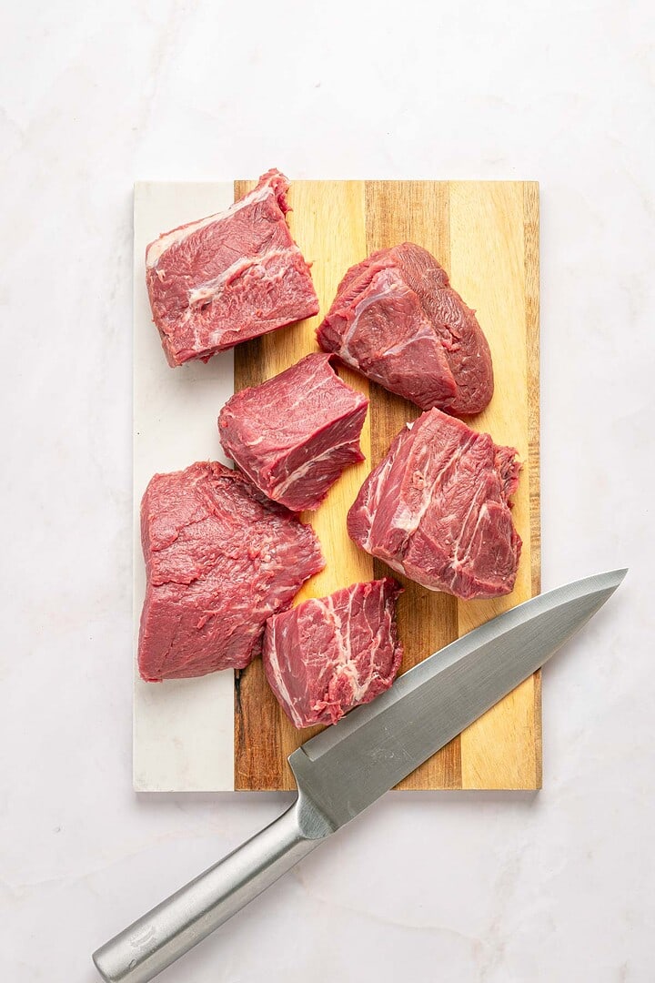 Six raw beef chunks on a wooden cutting board with a large chef&rsquo;s knife beside them, ready to be transformed into flavorful Birria Tacos or slow cooker favorites.
