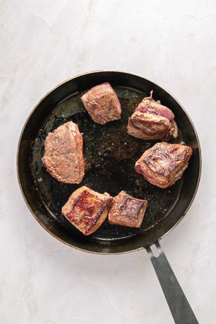 Five pieces of beef searing in a black skillet on a light-colored surface, perfect for making flavorful Birria Tacos.